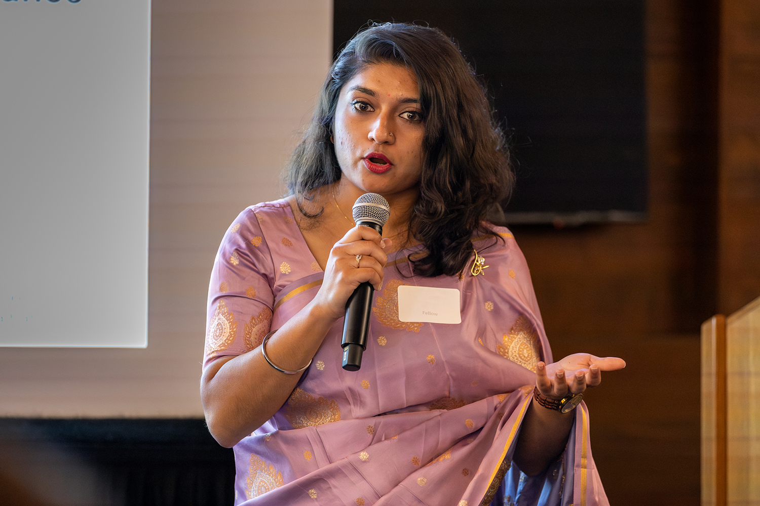 Karishma Bhagani holding a mic and giving a talk; she is dressed in a pink sari with gold embroidery.