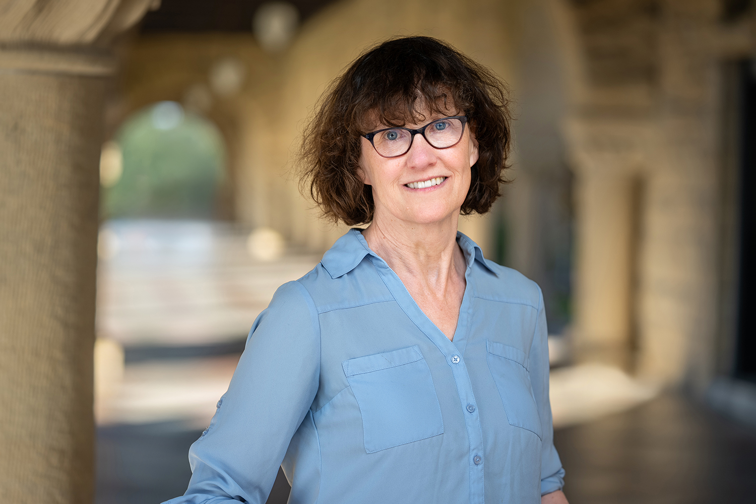 Krista Lawlor in a light blue shirt standing in a sunlit outdoor corridor, surrounded by arched stone pillars.