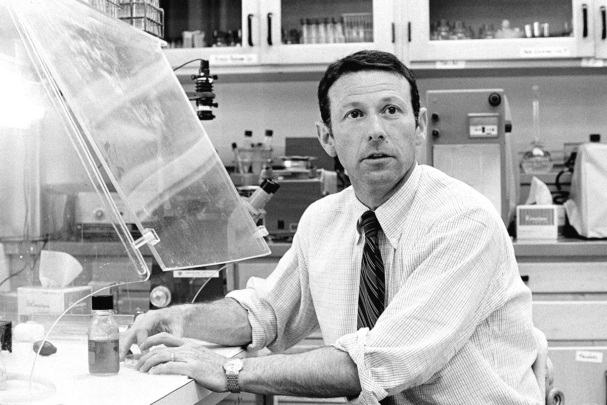 Black and white photo of Paul Berg in a shirt and tie sitting at a lab bench with equipment.