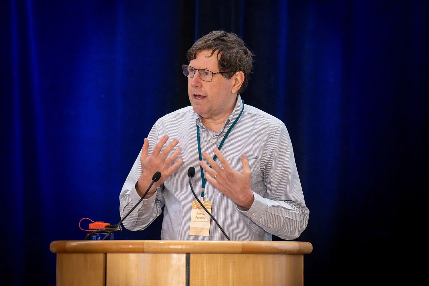 A man gestures while speaking at a podium during a conference, with a blue curtain in the background.