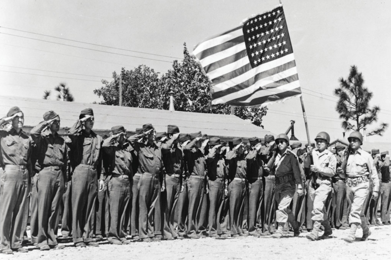 Black-and-white photo of a line of uniformed soldiers saluting as fellow soldiers march past carrying an American flag.