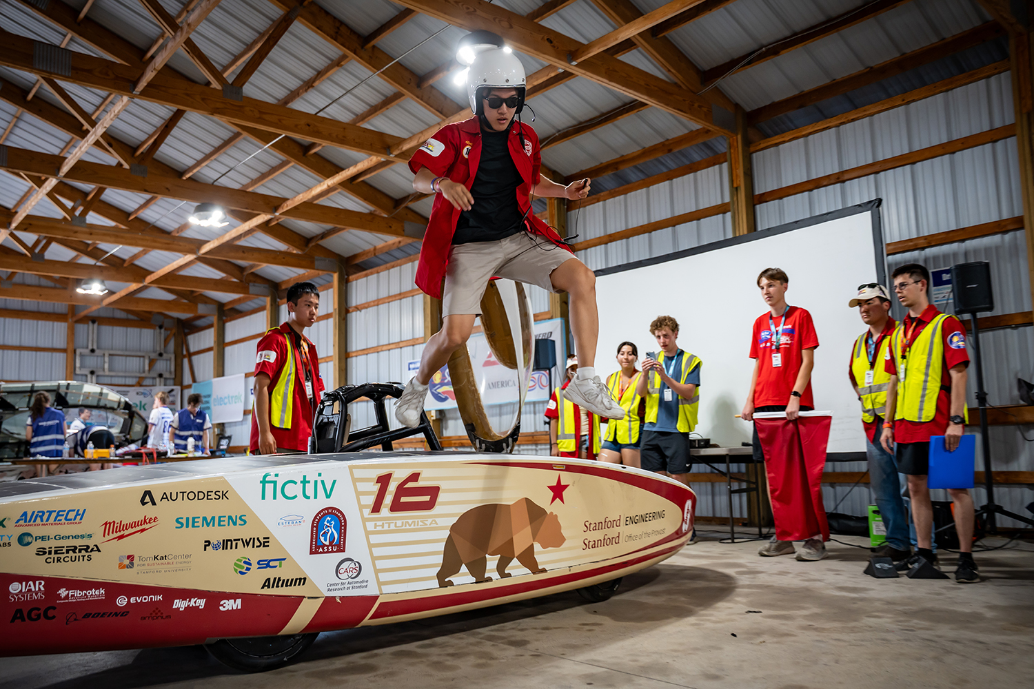 A helmeted team member leaps out of Stanford's solar car inside a warehouse with other team members in the background.
