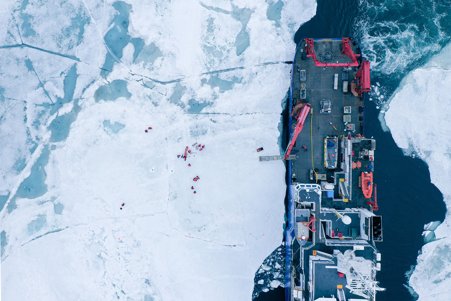 Aerial view shows a research ship breaking through Arctic sea ice with scientists working on the surface.