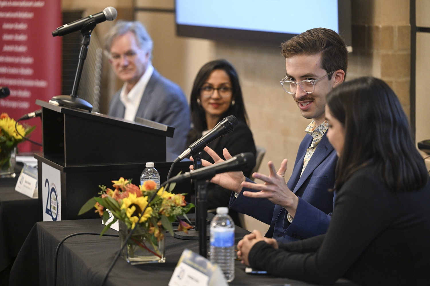 A panel discussion with participants seated at a table, microphones in front of them, and flowers as centerpieces.