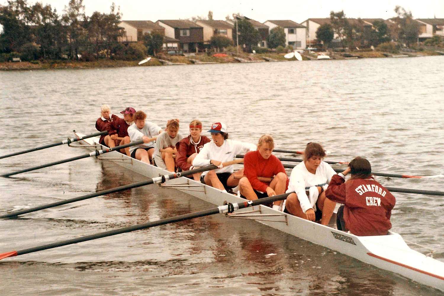 Kristin Smith and the Cardinal rowing team sit in their shell on a calm waterway, preparing for practice, with houses visible in the background.