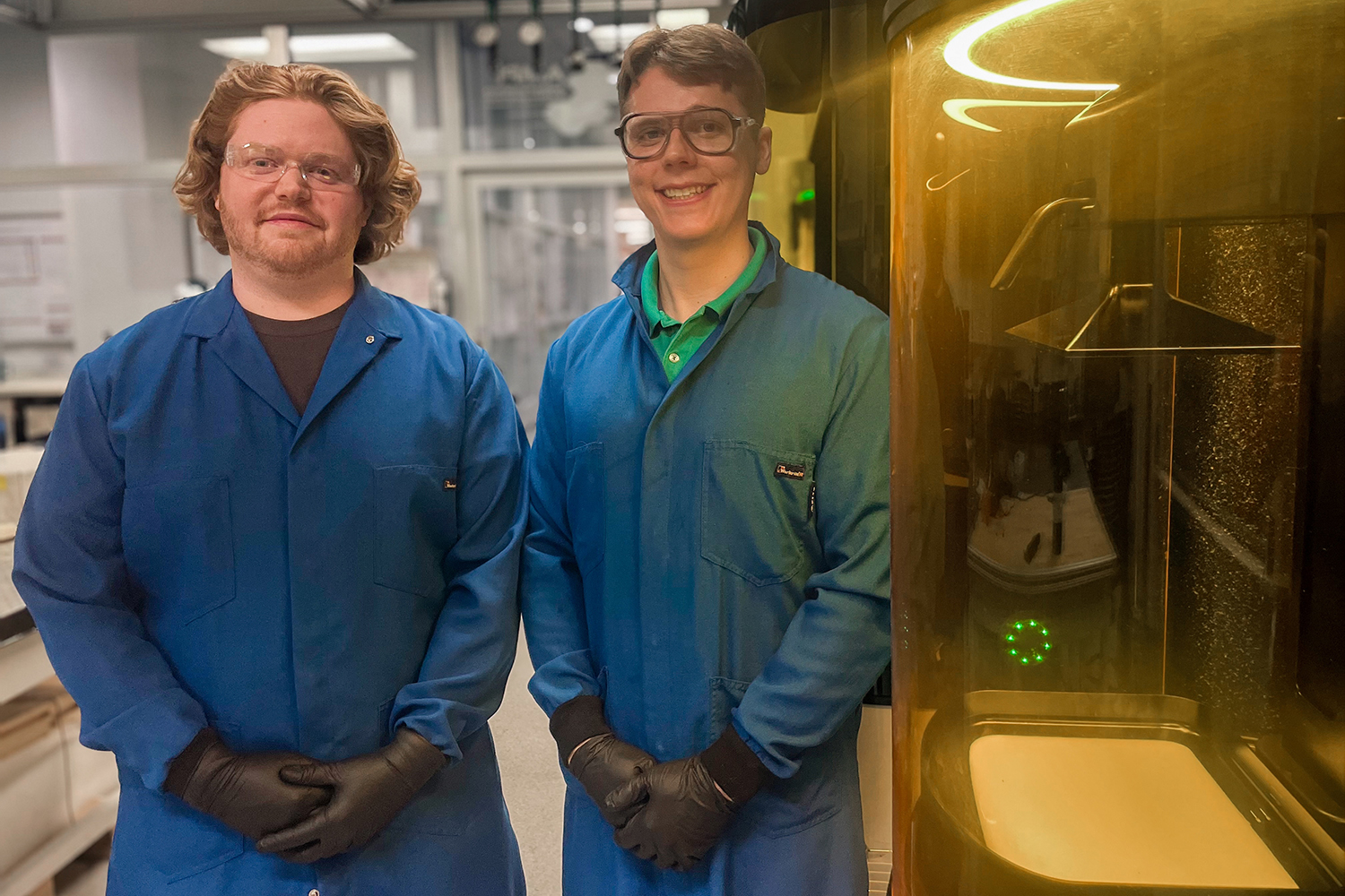 Jacob Dobson and Philip Onffroy in blue lab coats and gloves stand beside a 3D printer within a laboratory setting.