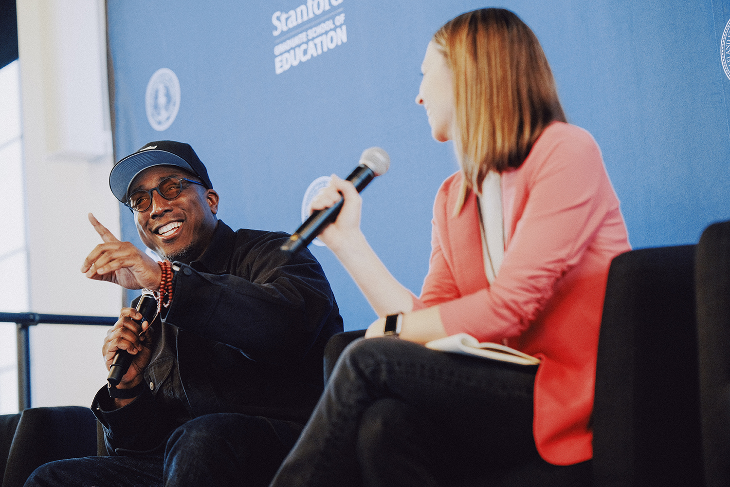 Kareem Edouard gestures while discussing a topic on stage, joined by a moderator holding a microphone.