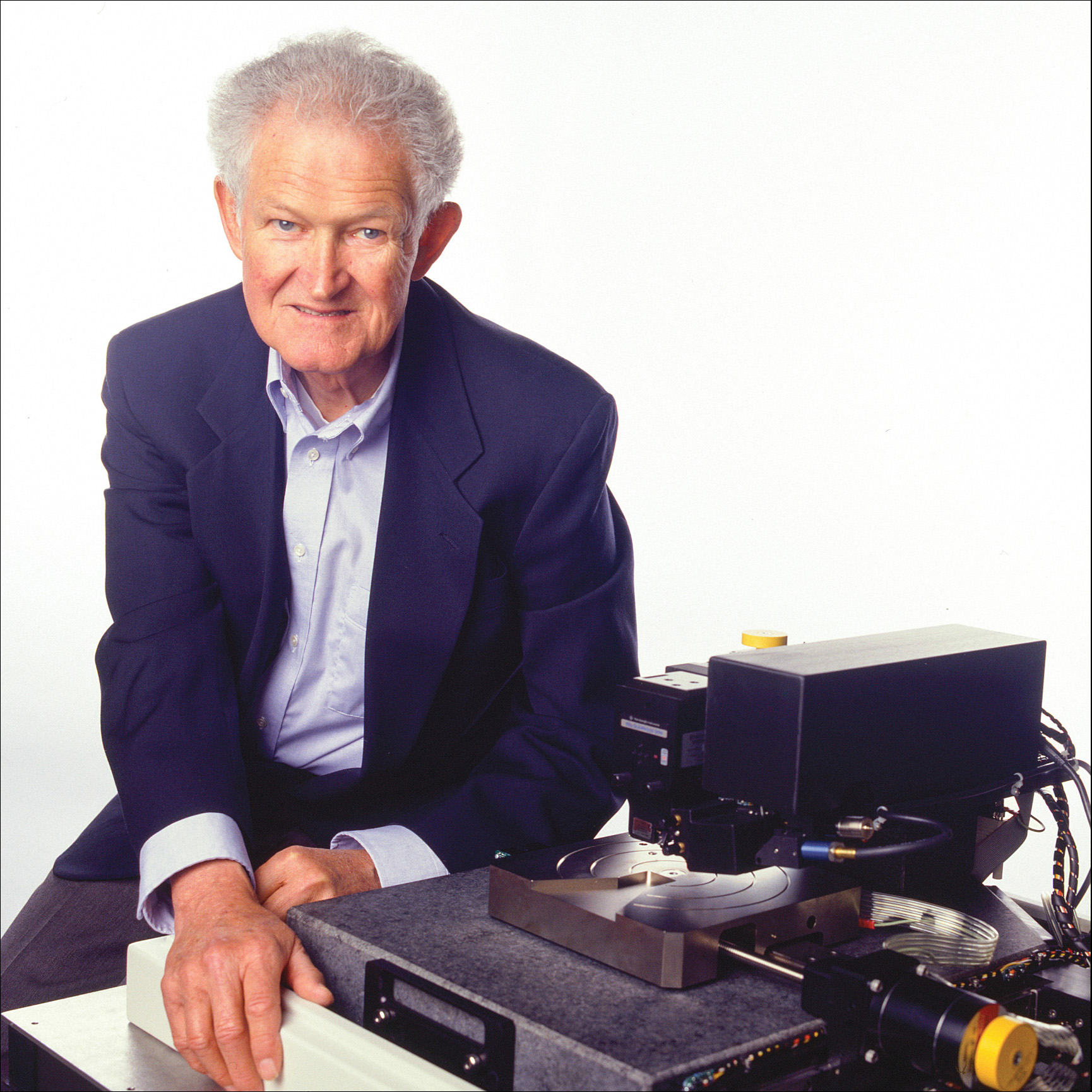 Calvin Quate in a navy suit sitting at a table with an atomic force microscope.