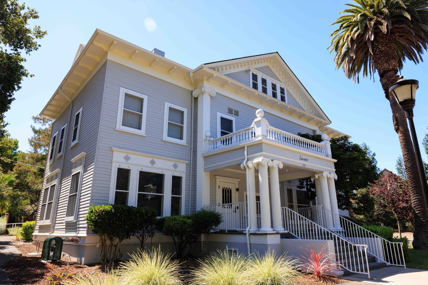 A light gray historic house features white columns, a front porch, and palm trees in the garden.