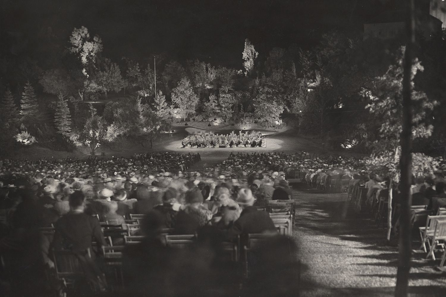 A crowd gathers at a nighttime outdoor event, seated in rows facing a stage with lights and trees surrounding the area.