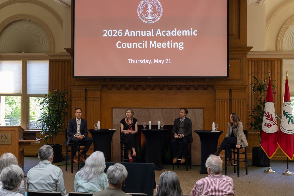 Four speakers participate in the 2026 Annual Academic Council Meeting, seated at black tables with an Audience in the foreground.