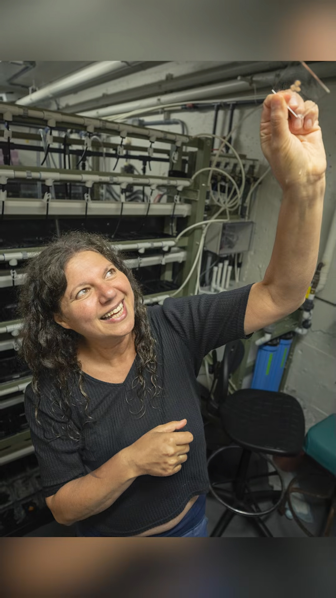 Ayelet Voskoboynik in a black shirt holds a scientific sample while aboard a research vessel, surrounded by equipment and machinery.