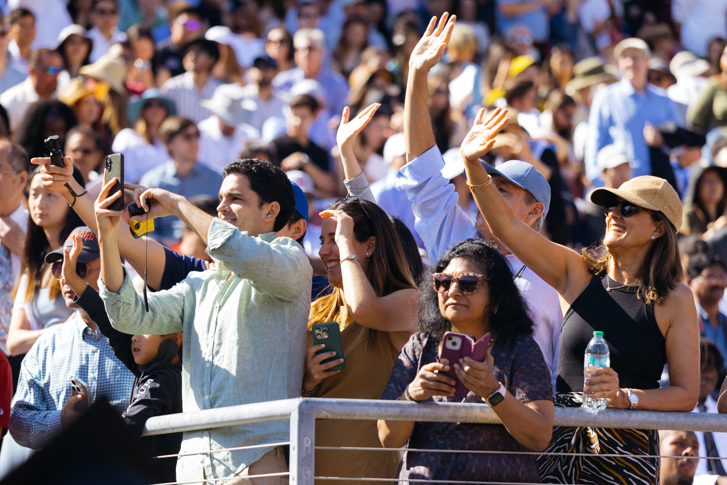 Spectators wave and take photos in crowded stadium stands during sunny commencement event.