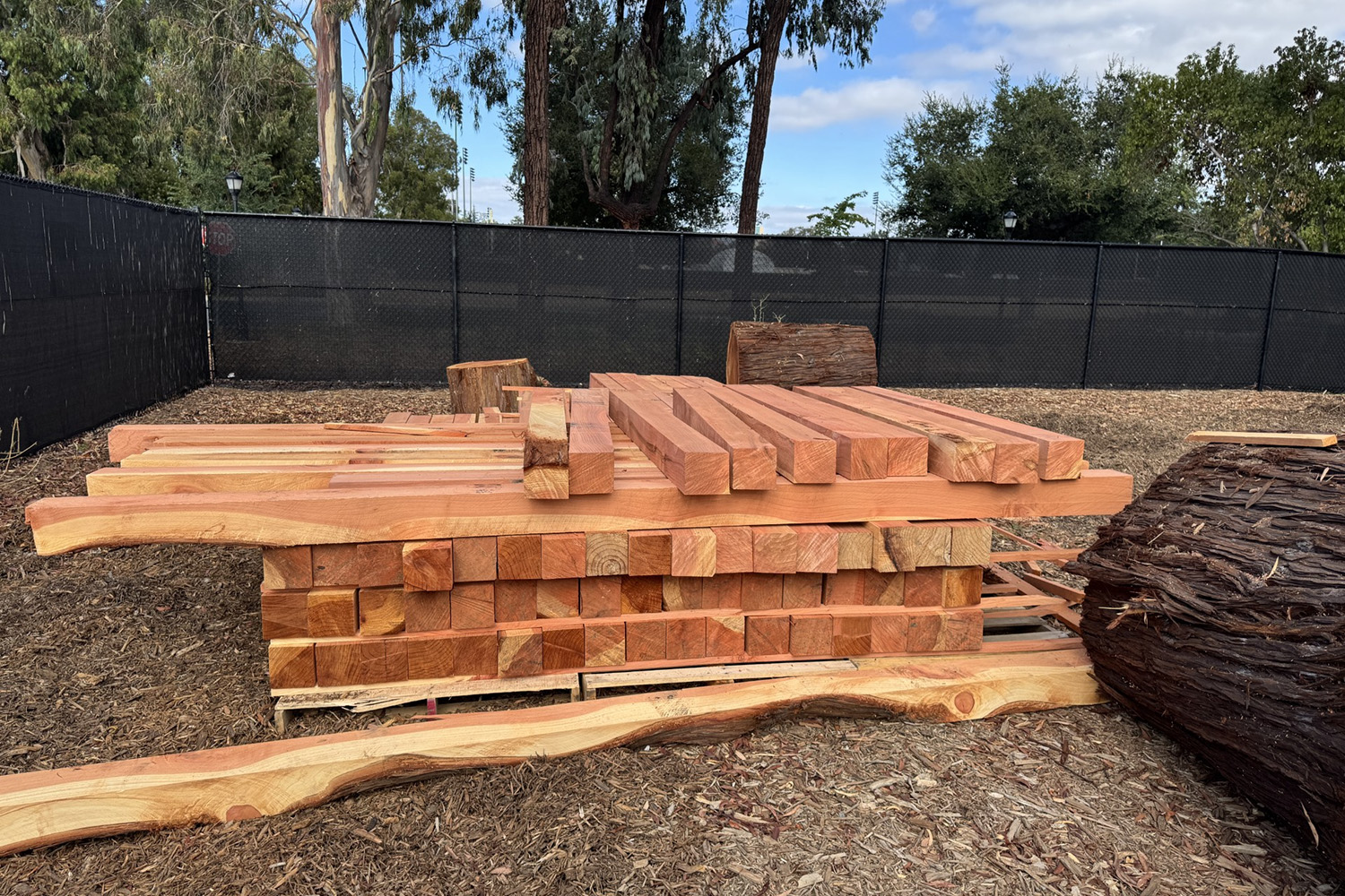 Wooden planks and beams are stacked on a wooden pallet in a fenced outdoor area.