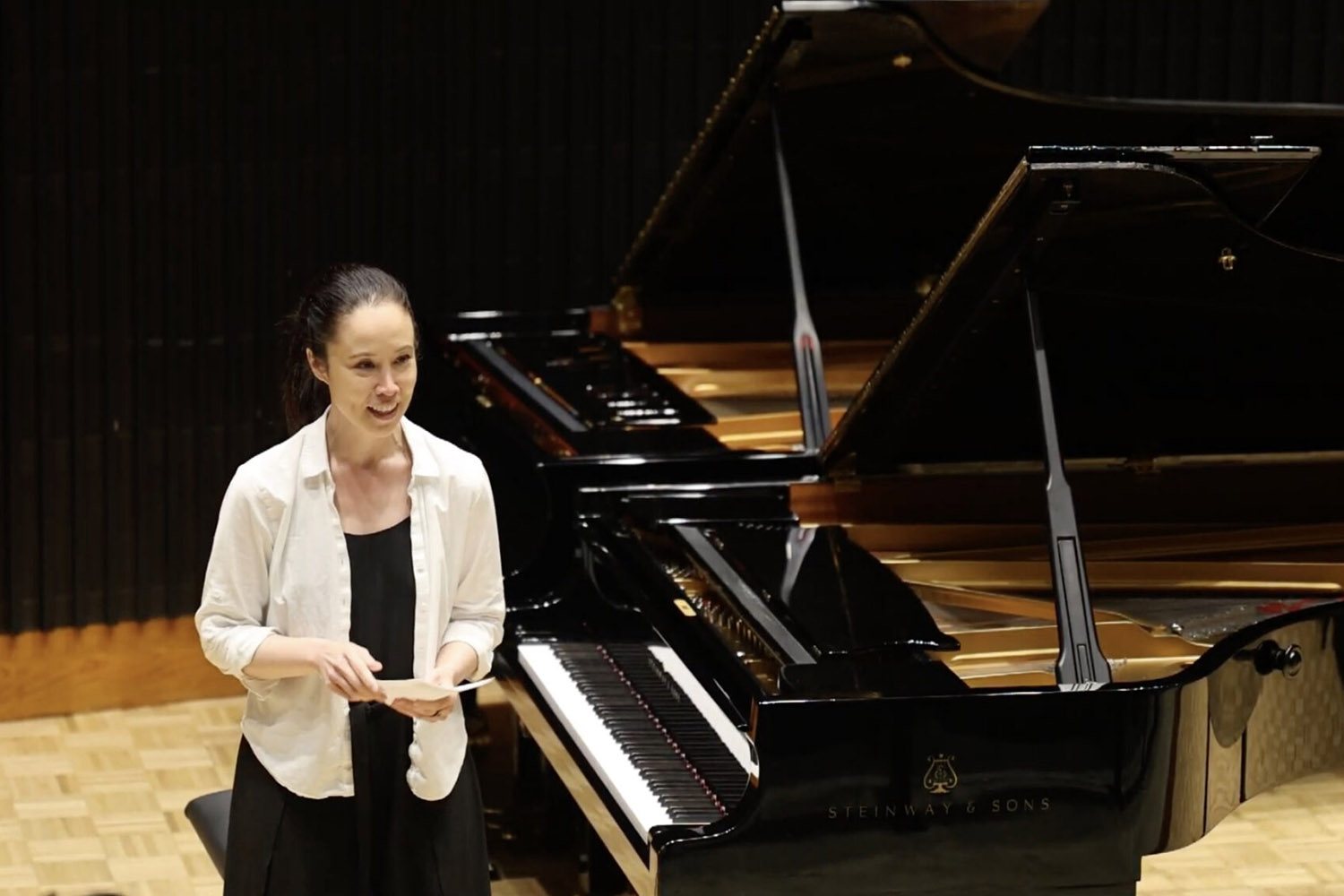 A woman stands by a Steinway piano, speaking to an audience in a performance setting.