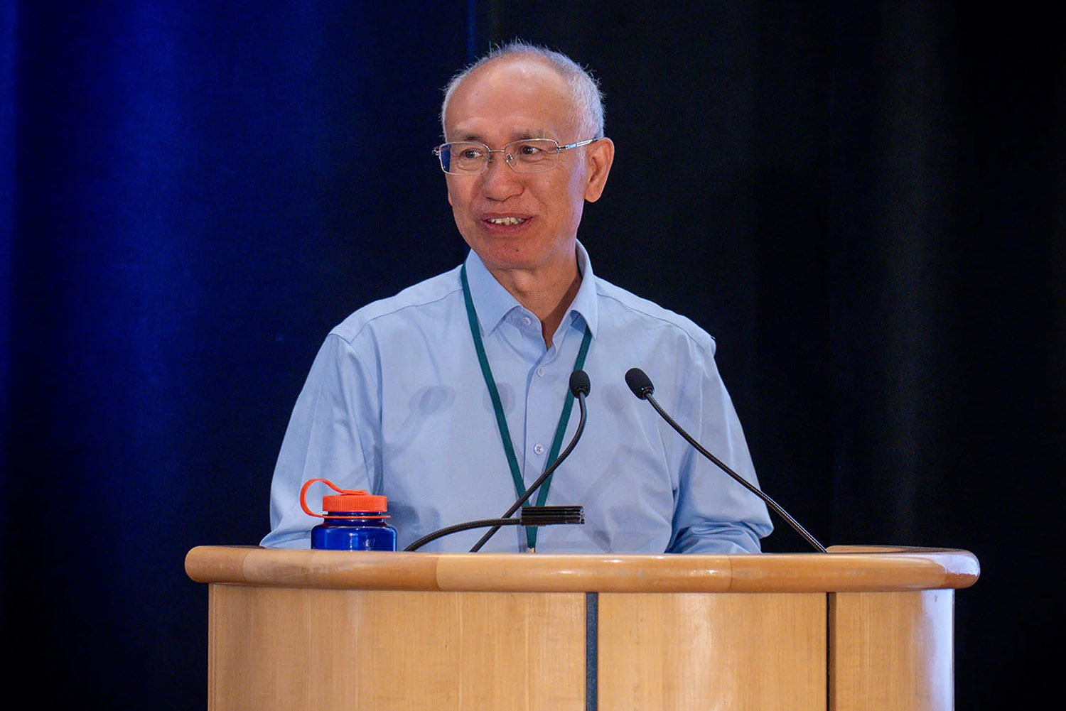 A man speaks at a podium during a presentation, with a water bottle and microphone visible in front of him.