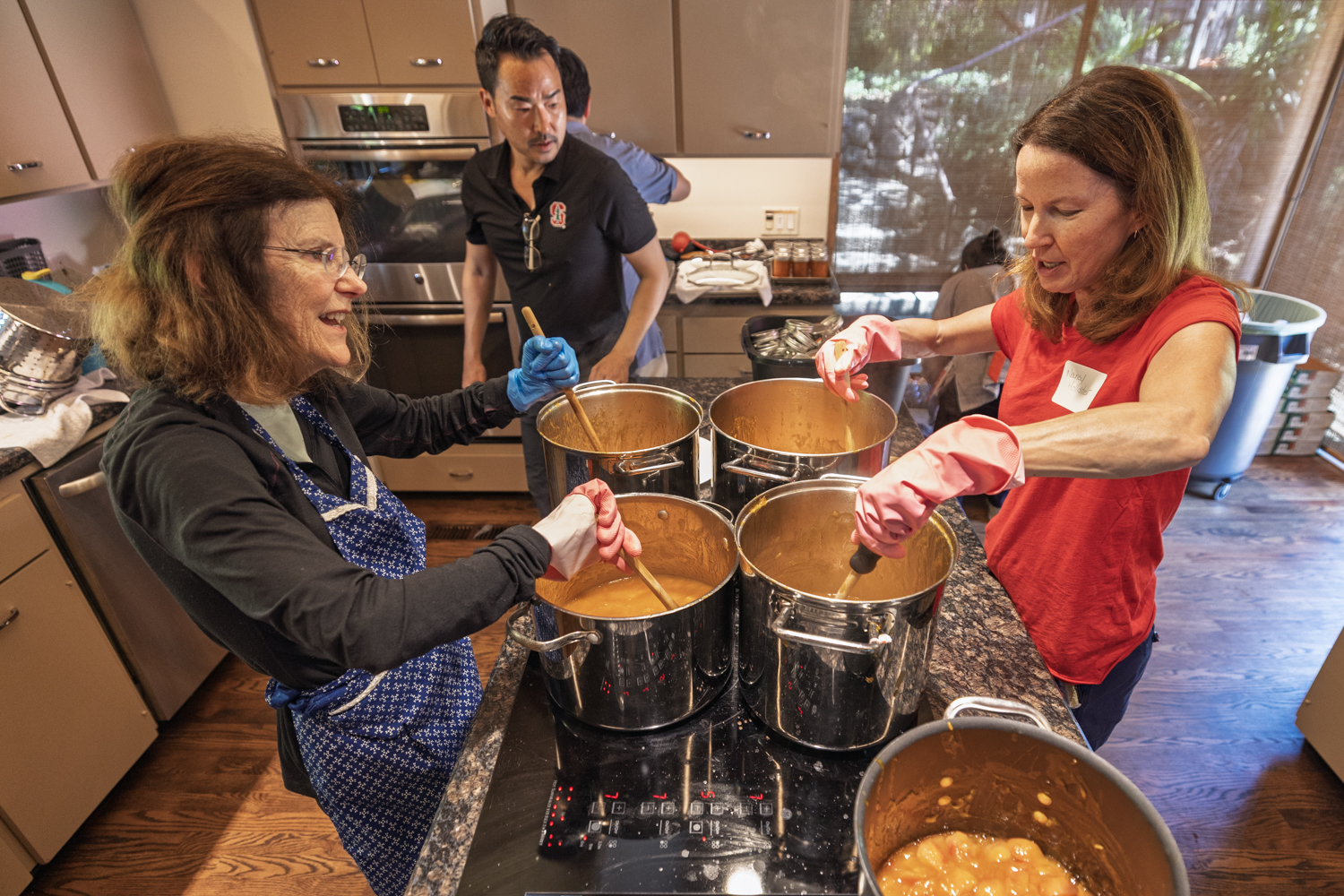 Group of people stirring large pots of apricot jam on a stovetop in a home kitchen.