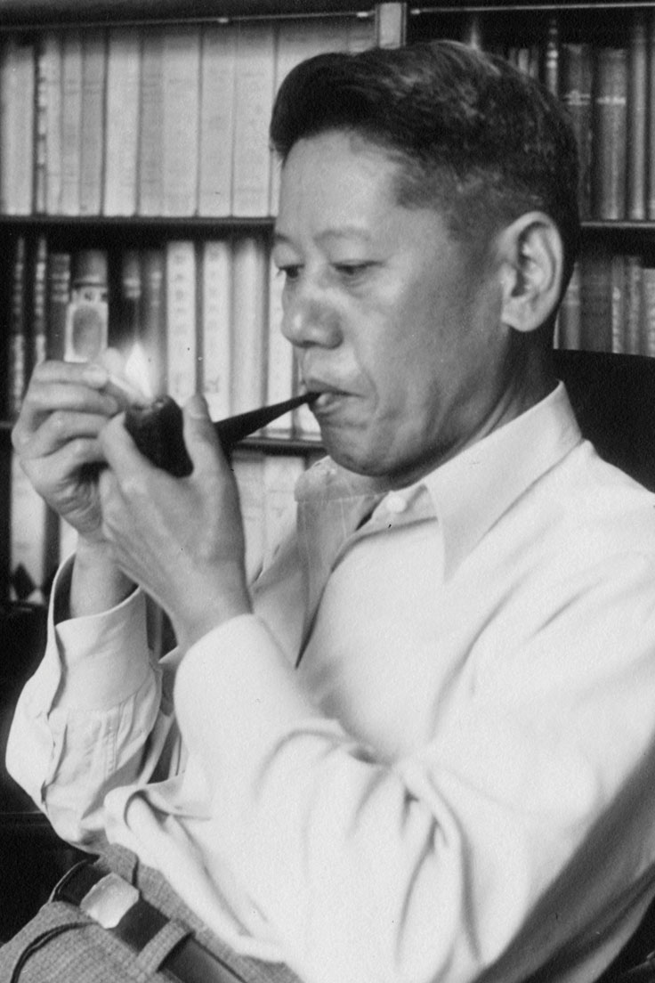 Black-and-white photo of Yamato Ichihashi lighting a pipe while seated in front of a bookshelf.