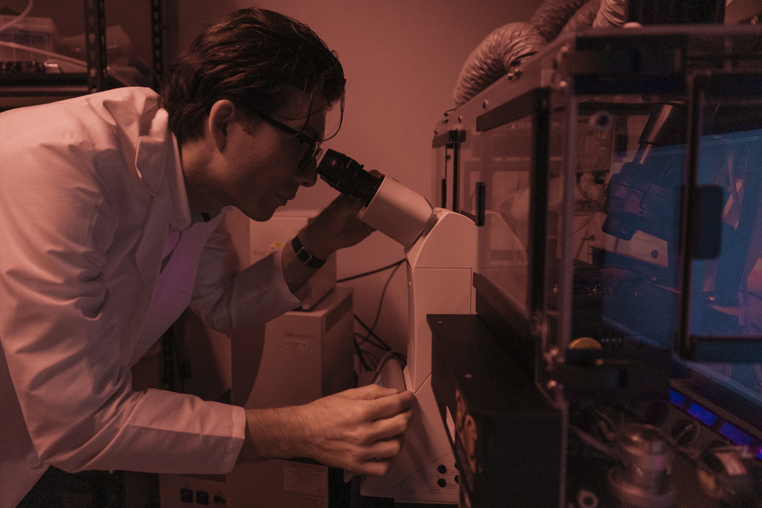 Sergiu Pasca in a white lab coat looks through a microscope in a dim, red-lit lab.