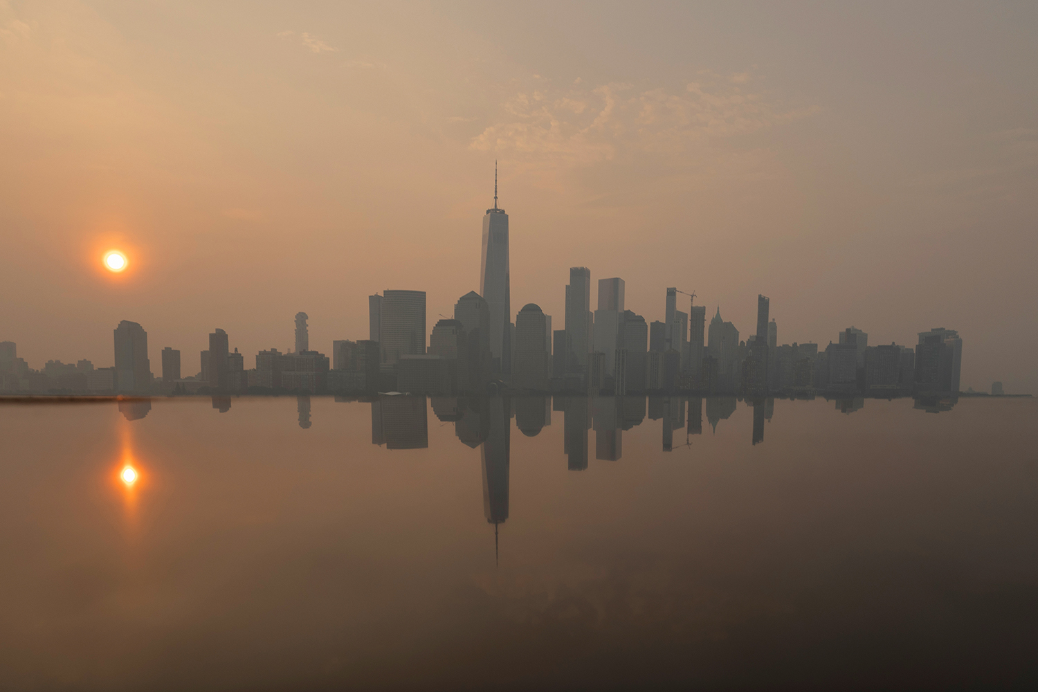 The Manhattan skyline sits under a haze at sunrise, its silhouette mirrored in the still water below.