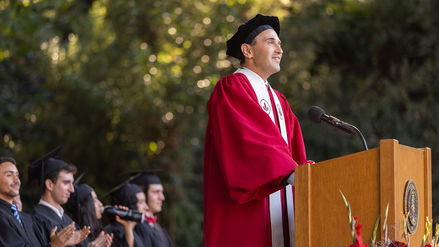 President Levin in crimson academic robe and black tam speaks at a podium while seated students in black gowns applaud.