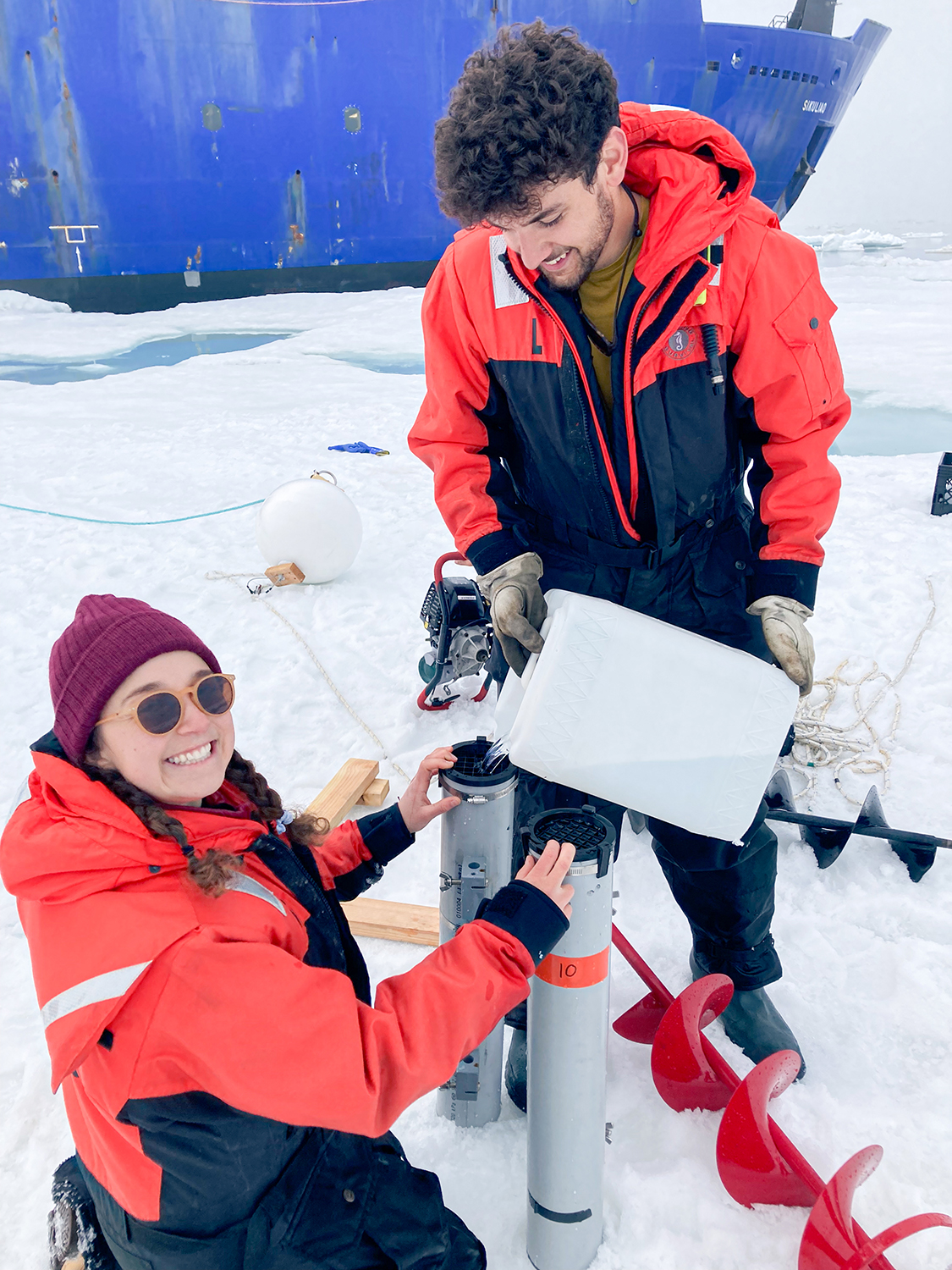 Two researchers in cold-weather gear work together to extract an ice core sample near the ship.