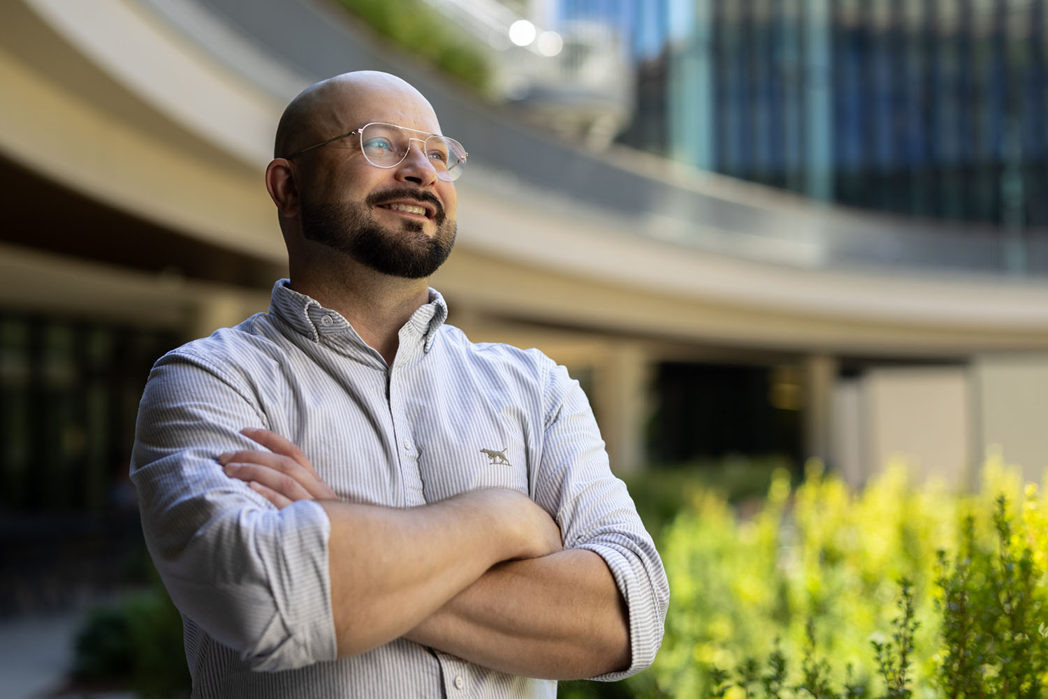 Mitchell Peterson, bearded and wearing glasses and a striped shirt, stands outdoors with arms crossed, smiling upward.
