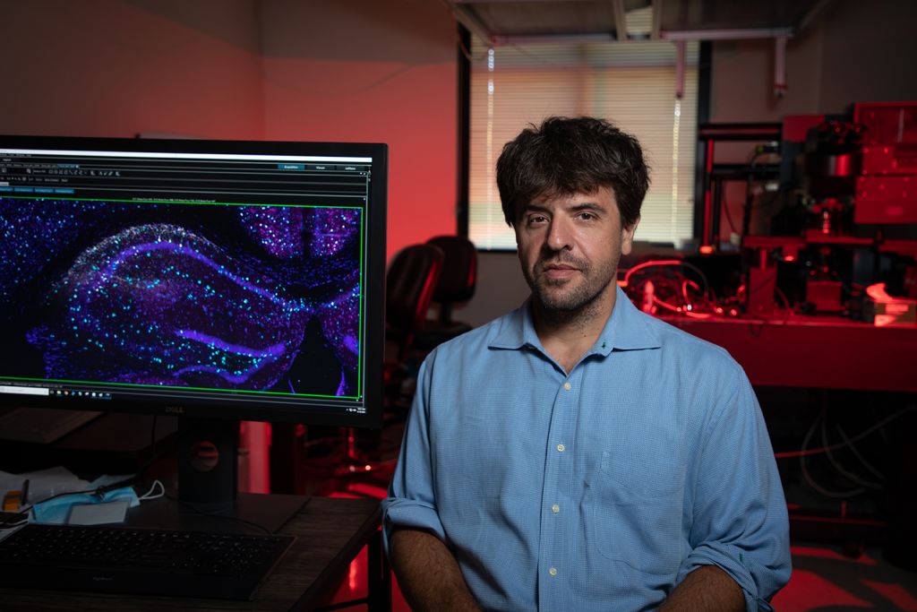 Karl Deisseroth sits in a red-lit lab beside a monitor showing a purple and teal microscope image.