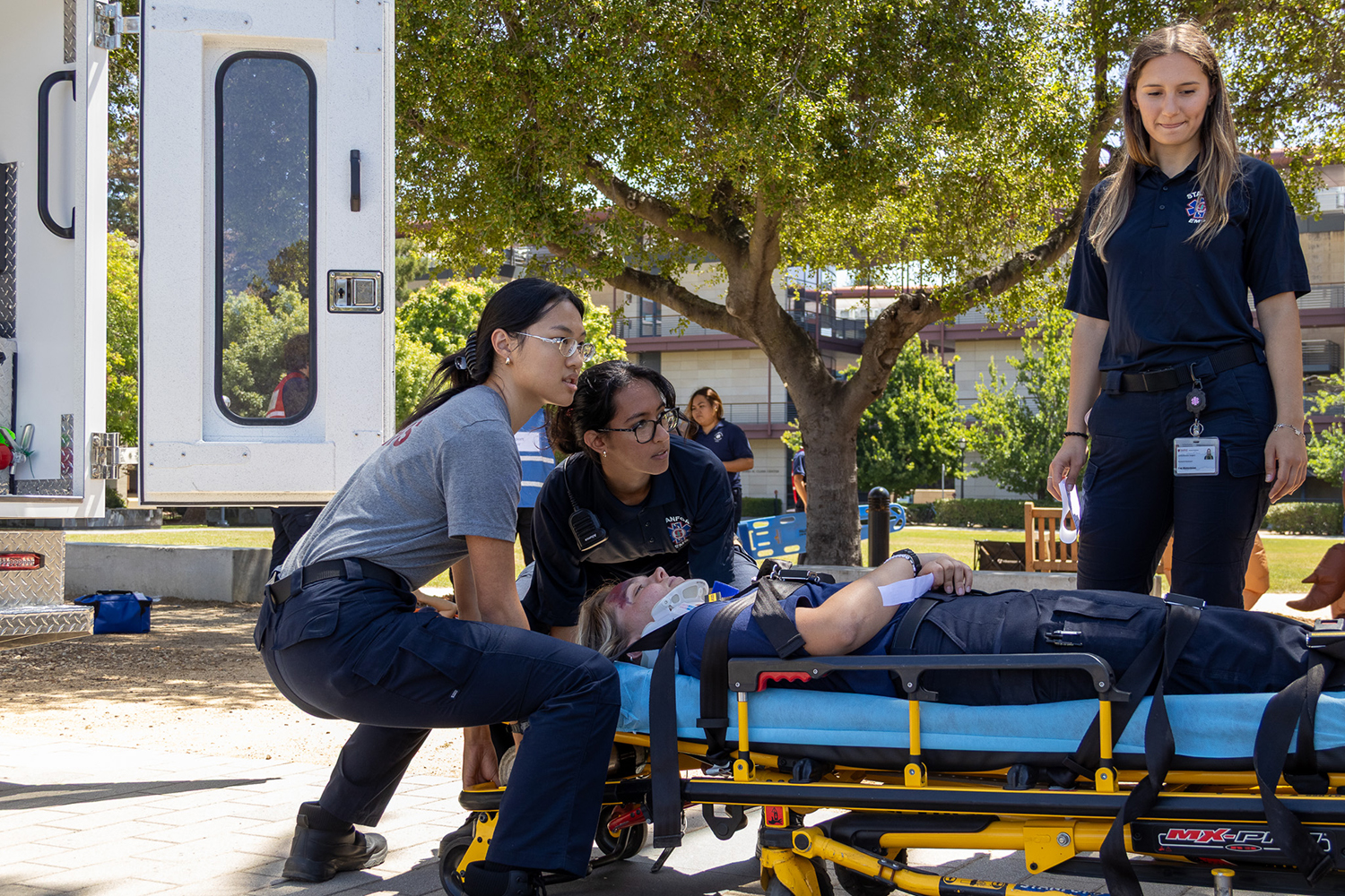 Student emergency responders practice on a stretcher beside an open ambulance door while a teammate looks on, during a training exercise outdoors.