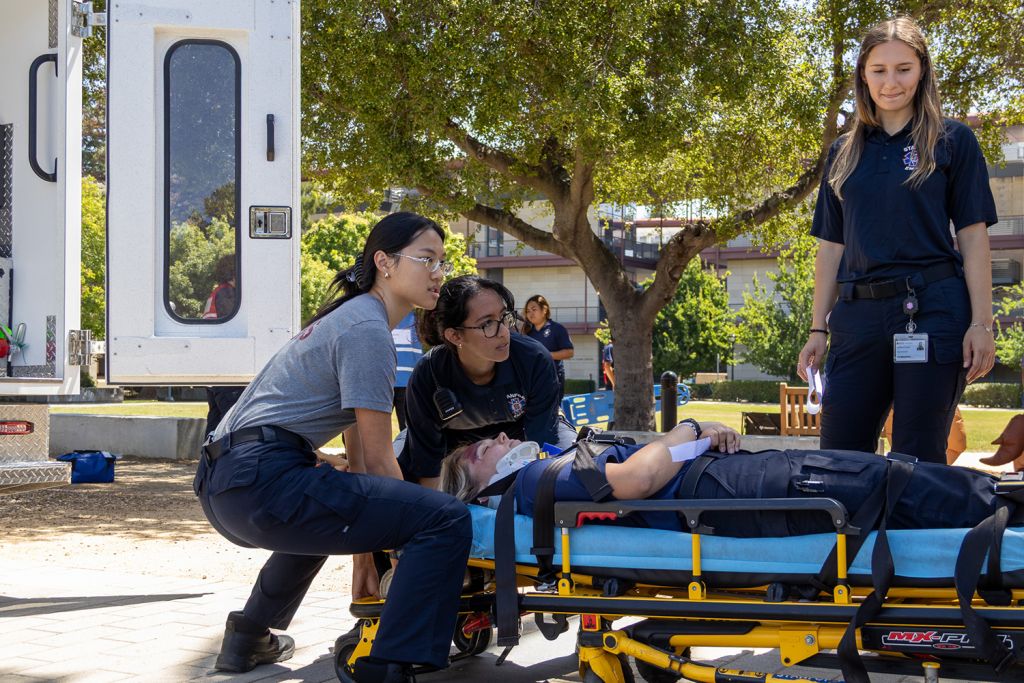 Student emergency responders practice on a stretcher beside an open ambulance door while a teammate looks on, during a training exercise outdoors.