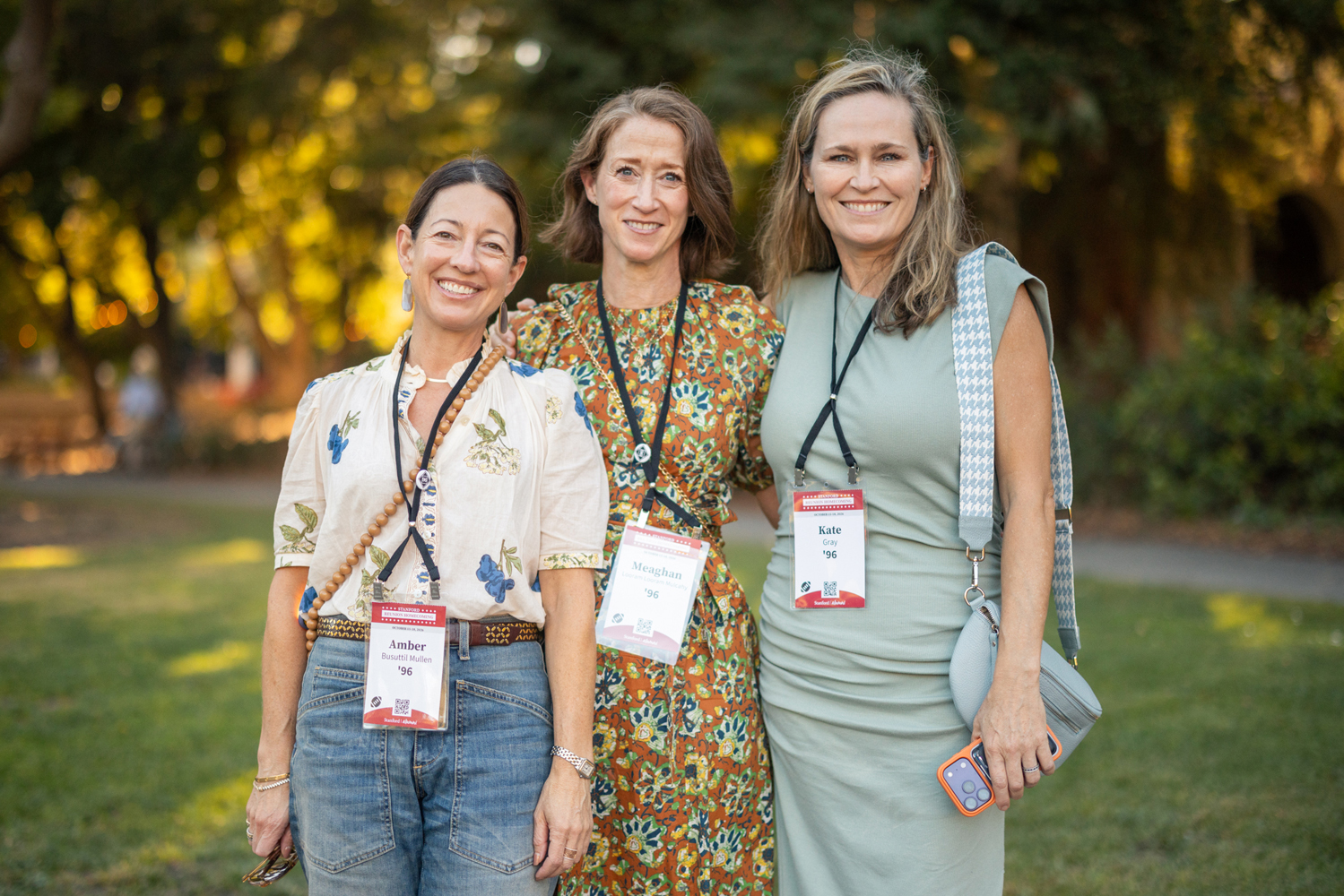 Three women smile together on a lawn in warm evening light, each wearing an event name badge.