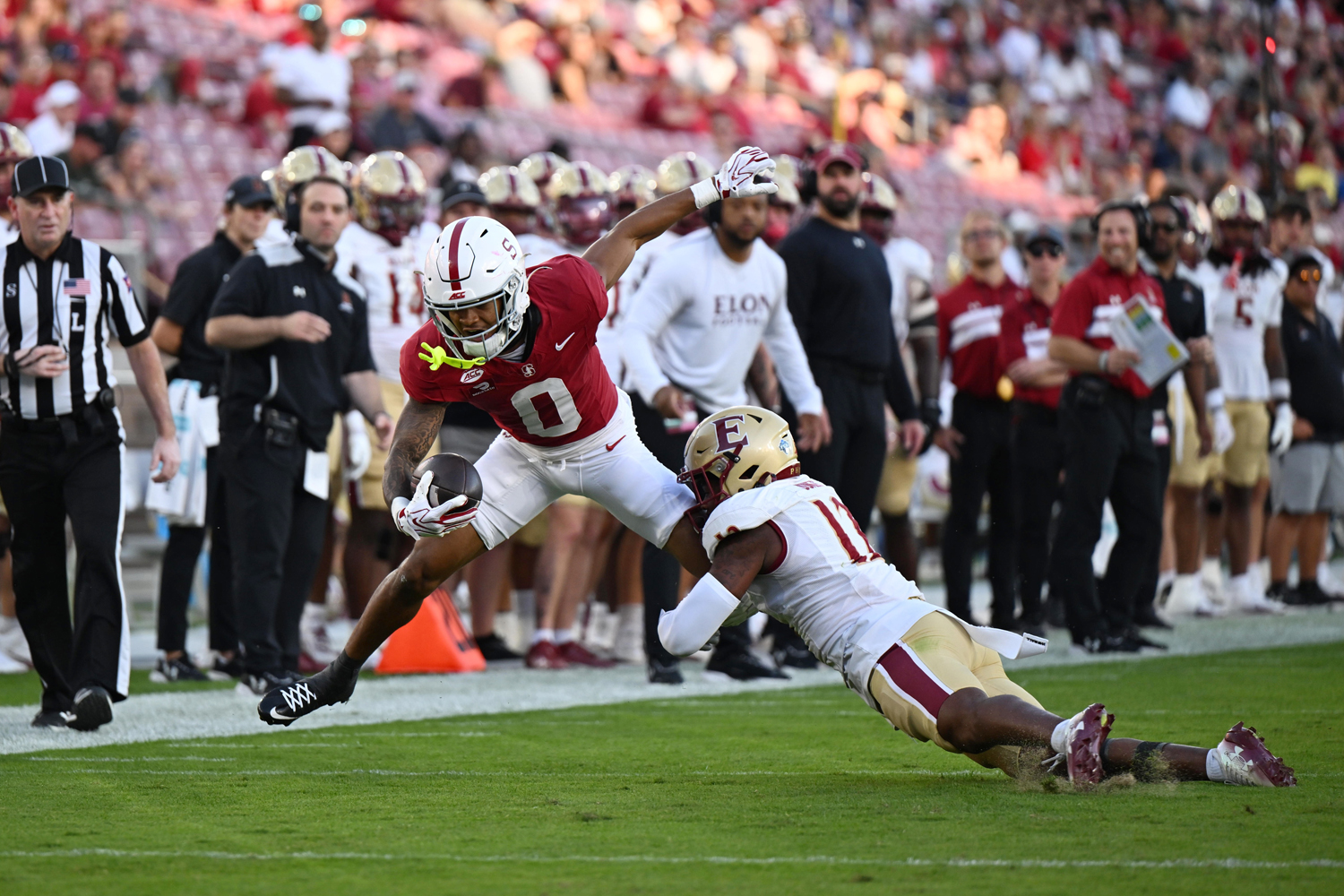 Player in a Cardinal uniform holds the football tight while a defender in white wraps him up near the sideline during a game, with coaches looking on.