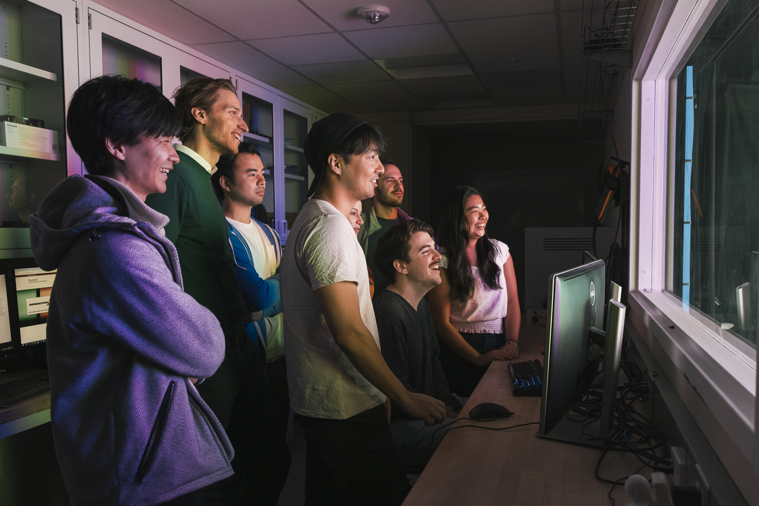 Group of researchers stand behind a computer, awash in a rainbow glow from lighting in the lab that's behind the computer, off-screen 