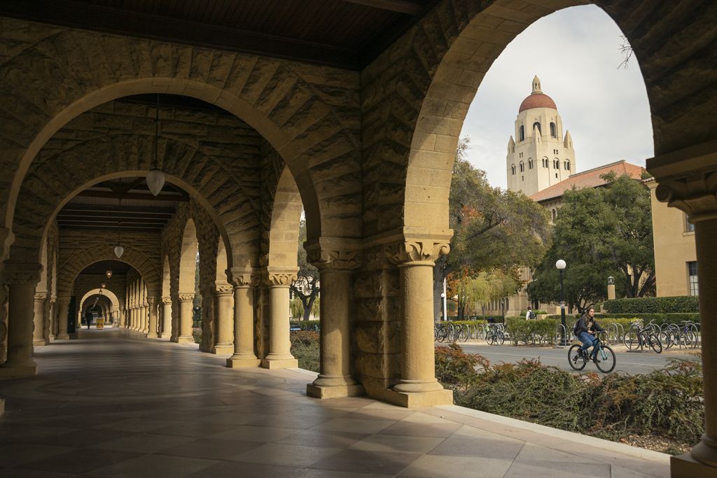 An arcade alternatingly in light and in shade as concentric arches let in fading sunlight. Hoover Tower is visible through the nearest arch.