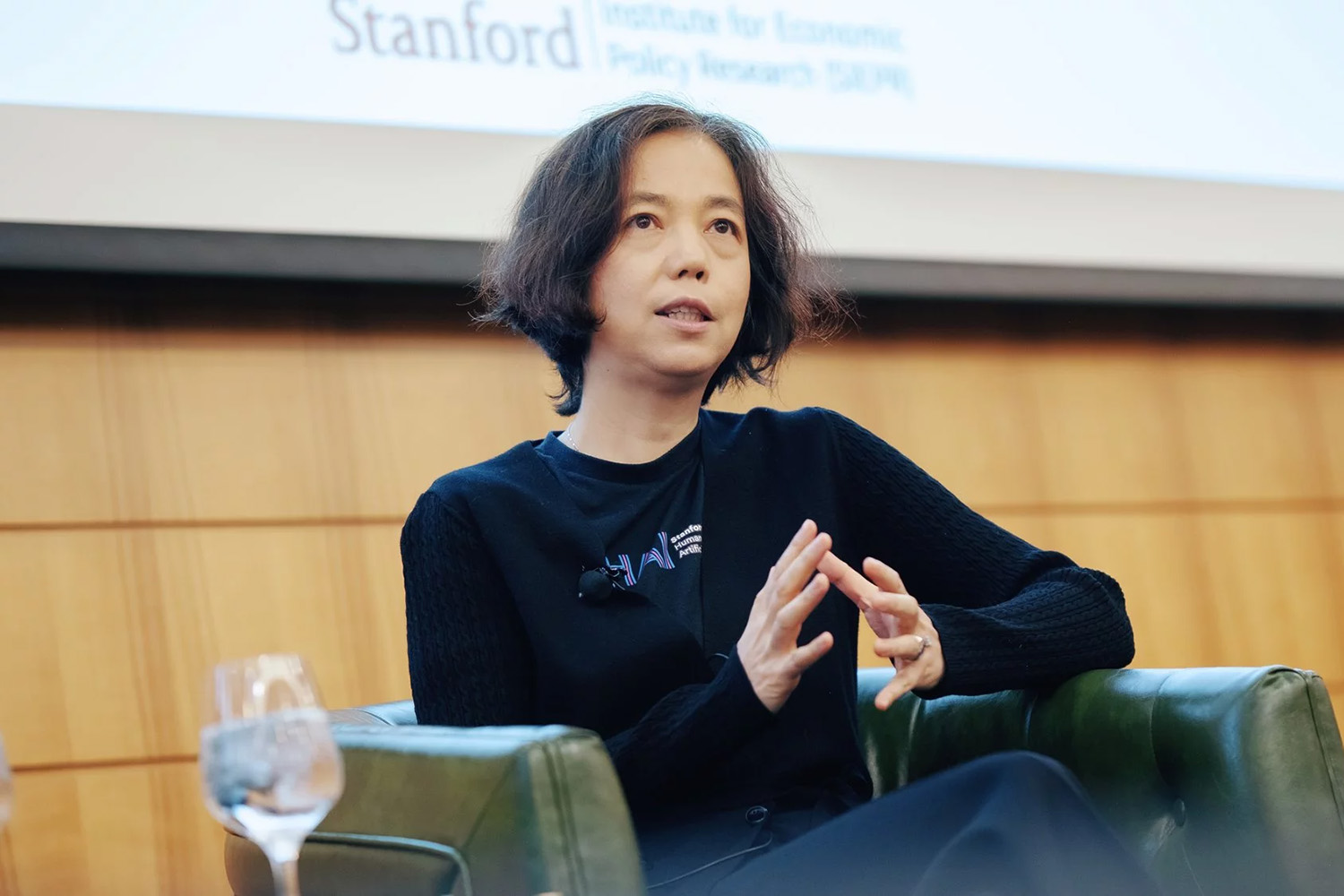 Fei-Fei Li, wearing a Stanford HAI sweatshirt, seated in a club chair onstage at the SIEPR Policy Forum.