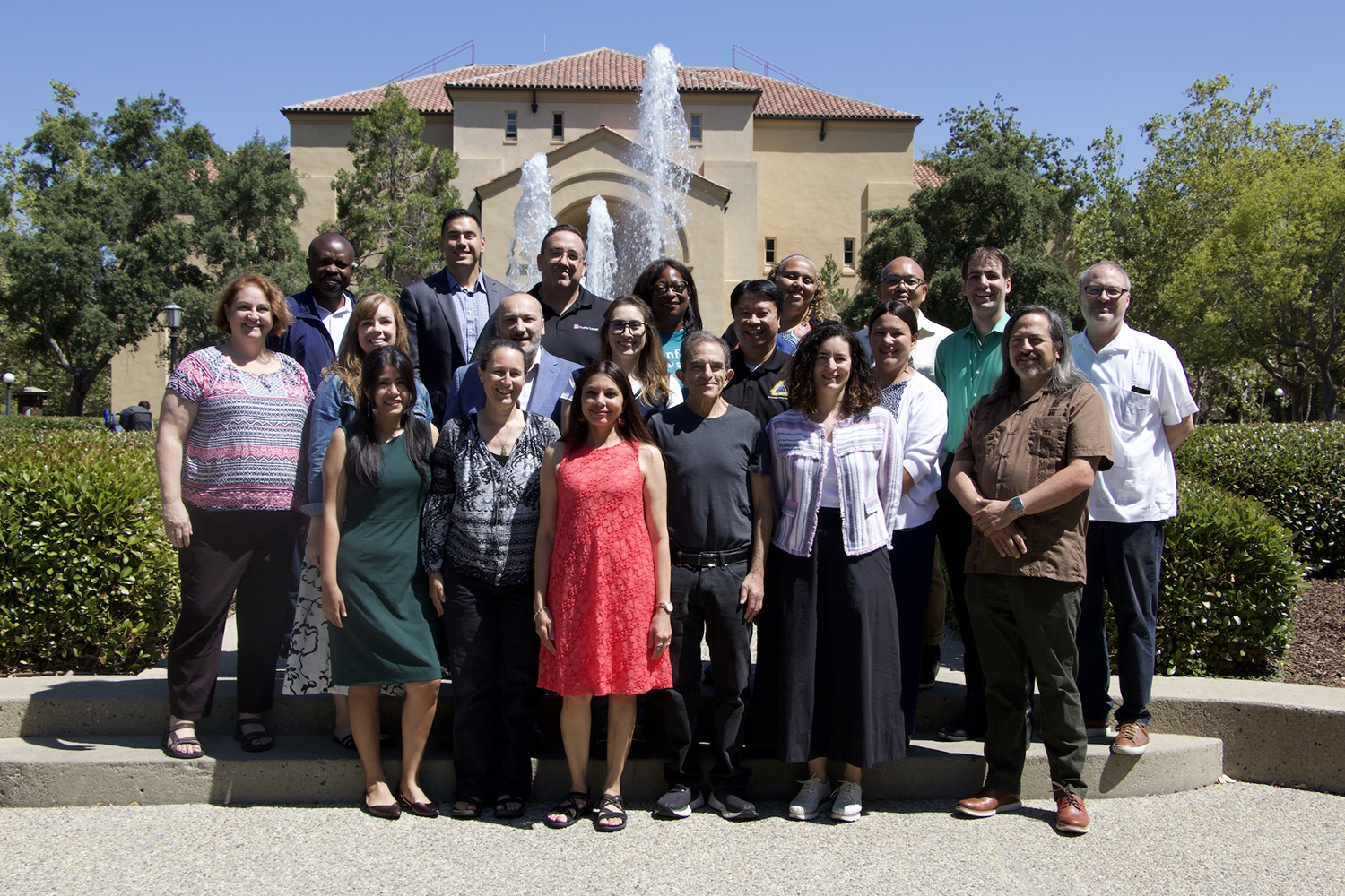 A group of 15 people stands together in front of a fountain and a building on a sunny day, smiling for the camera.