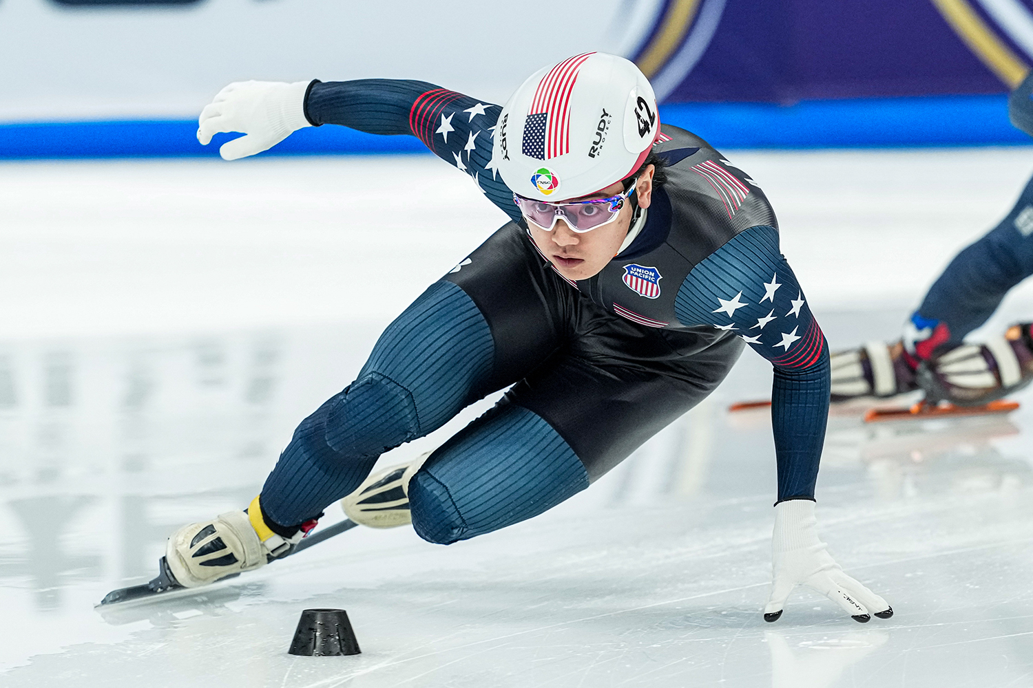 Brandon Kim wearing a form-fitting suit with American flag designs skates swiftly around a turn on an ice rink during a competition.