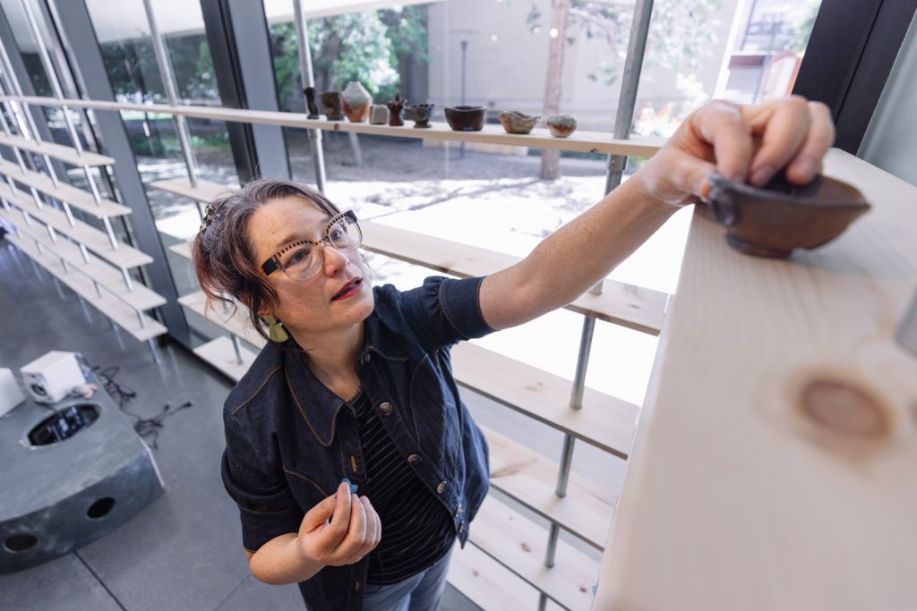 Open video Artist places a bowl on a high shelf in a bright studio with pottery displayed on shelves in the background. in a modal