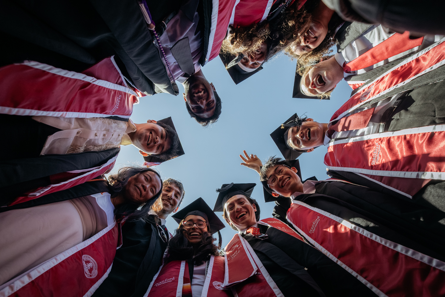 Graduates in caps and gowns with red stoles smile in a circle, photographed from below.