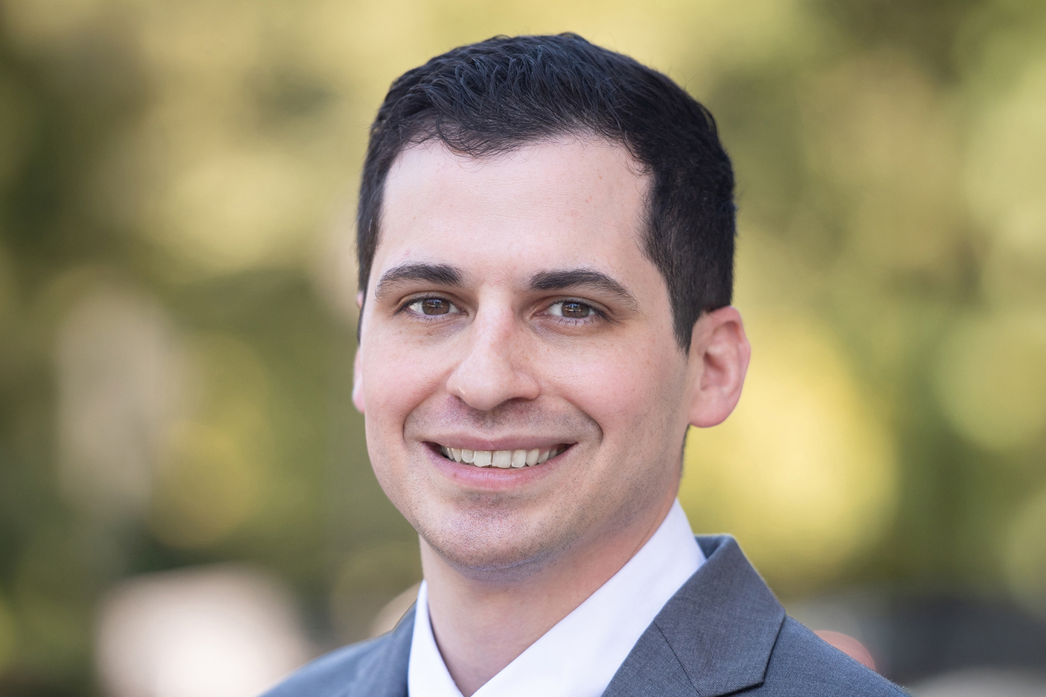 Daniel Delitto, a smiling man in a gray suit, poses for a professional headshot with a blurred green outdoor background.