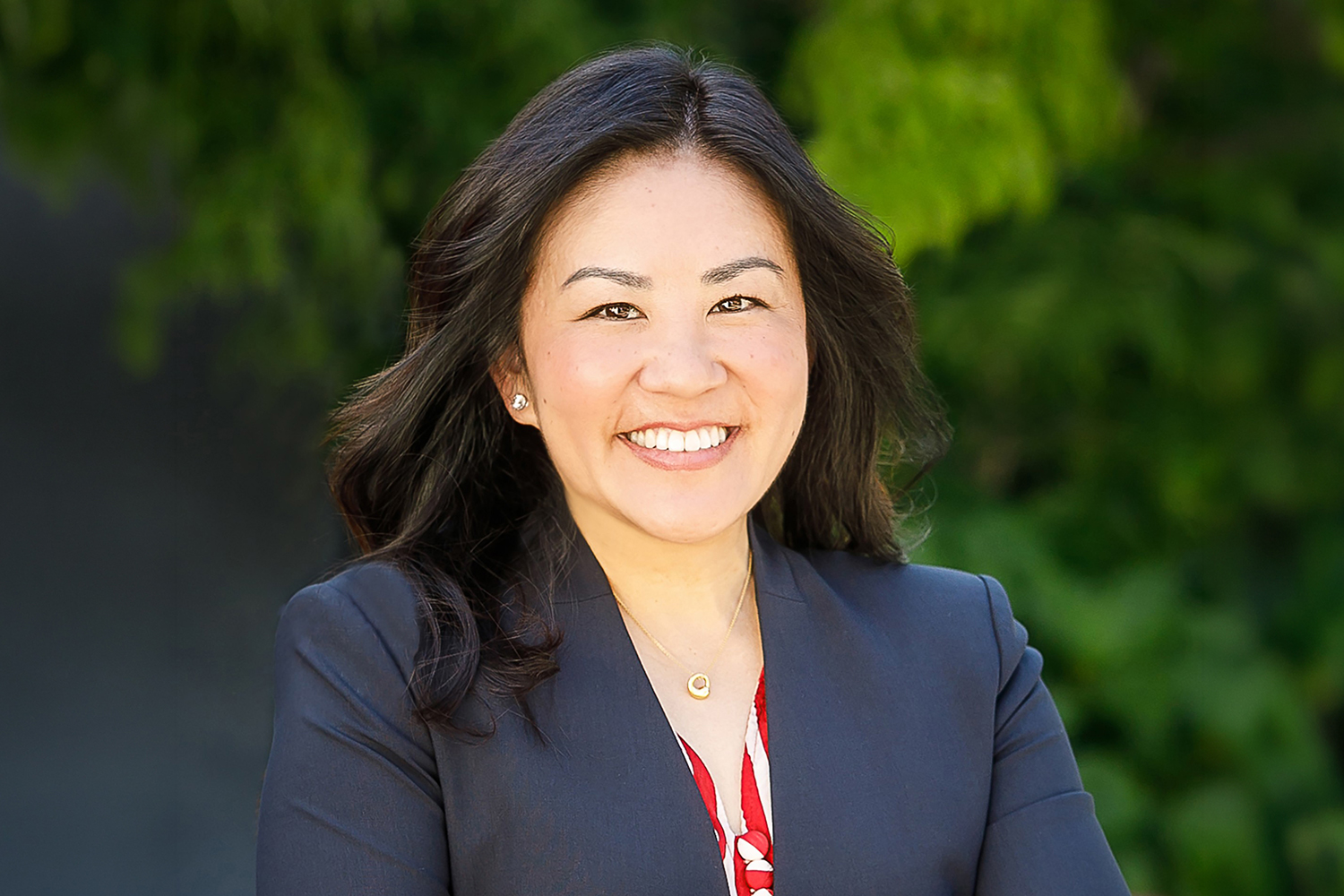 Smiling Reiko Osaki in a blazer stands outdoors with greenery in the background.