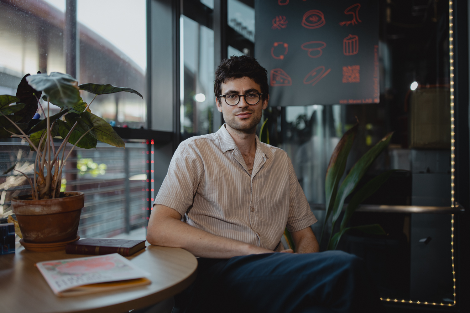 Vayu Hill-Maini sits at a lab cafe table beside a leafy potted plant, looking toward the camera.