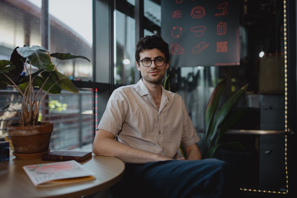 Vayu Hill-Maini sits at a lab cafe table beside a leafy potted plant, looking toward the camera.