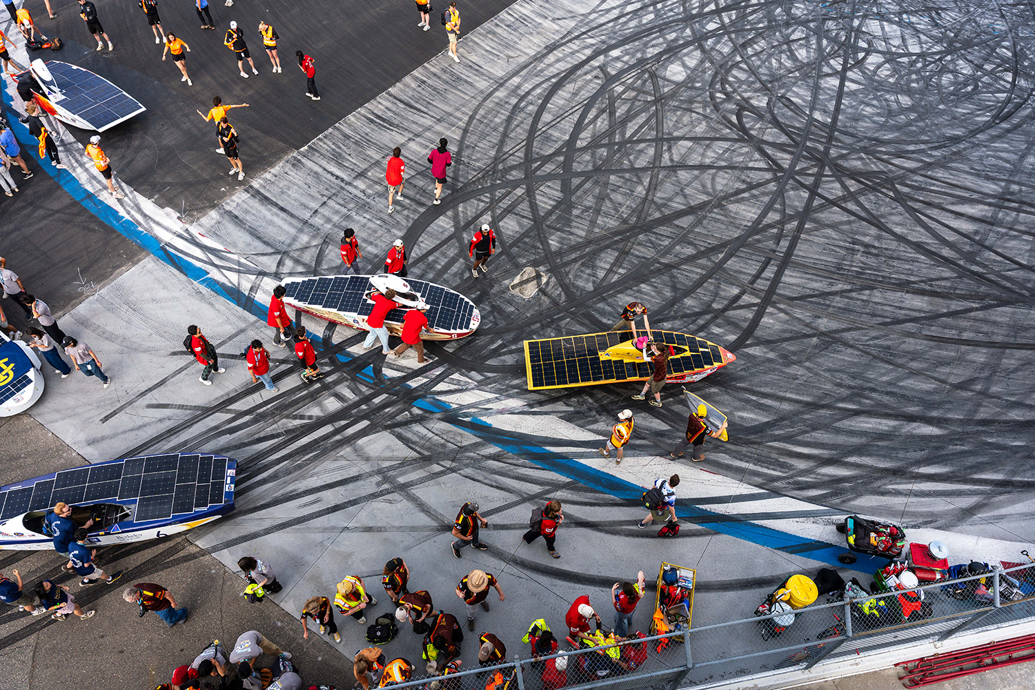 Aerial view of teams pushing their solar cars across a racetrack covered in swirling tire tracks.