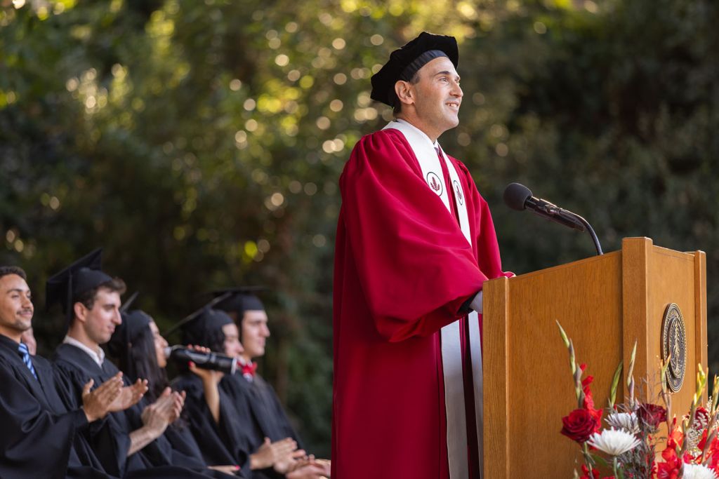 Open video President Levin in crimson academic robe and black tam speaks at a podium while seated students in black gowns applaud. in a modal