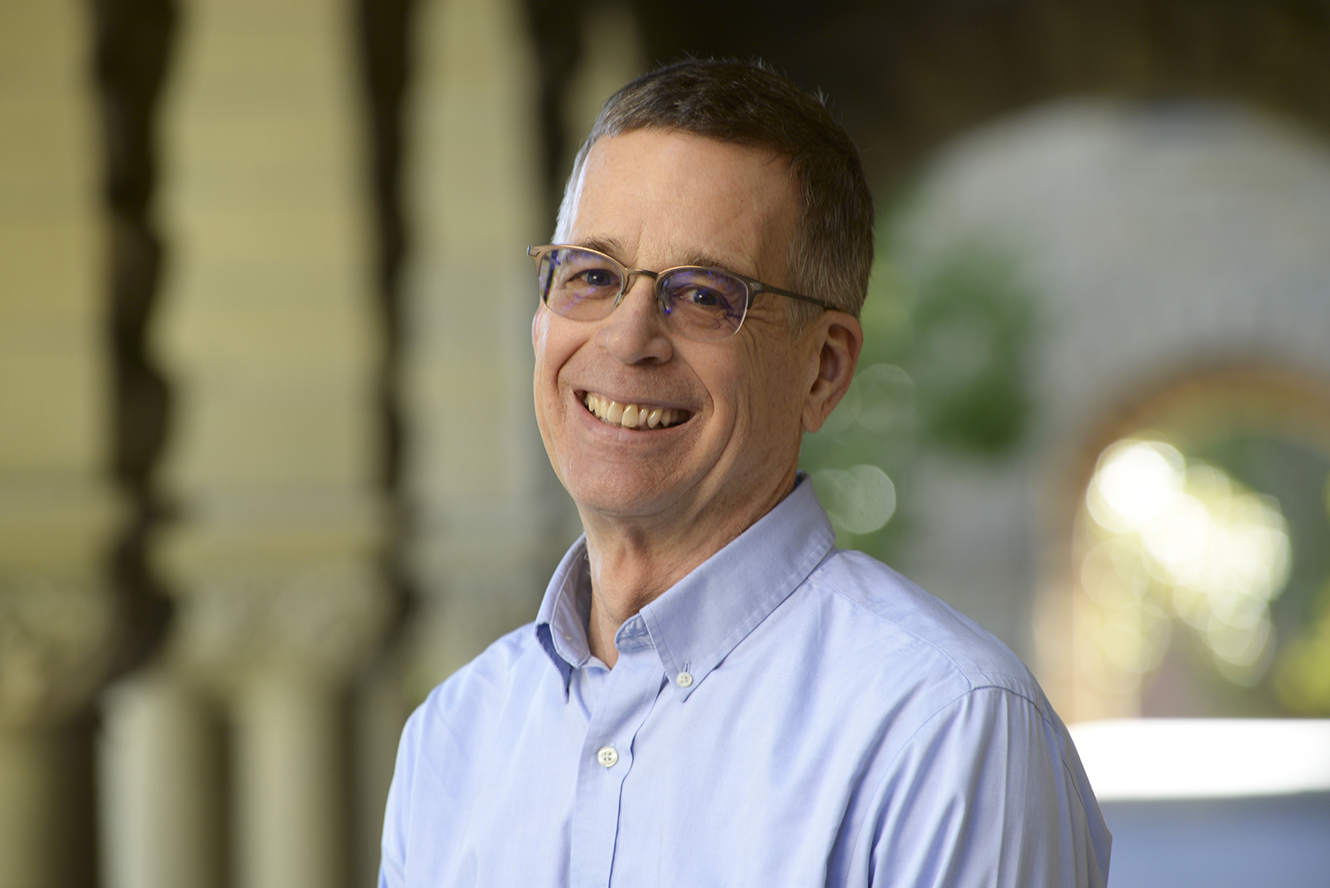 Steve Luby poses for a professional headshot outdoors with a blurred arched campus corridor in the background.