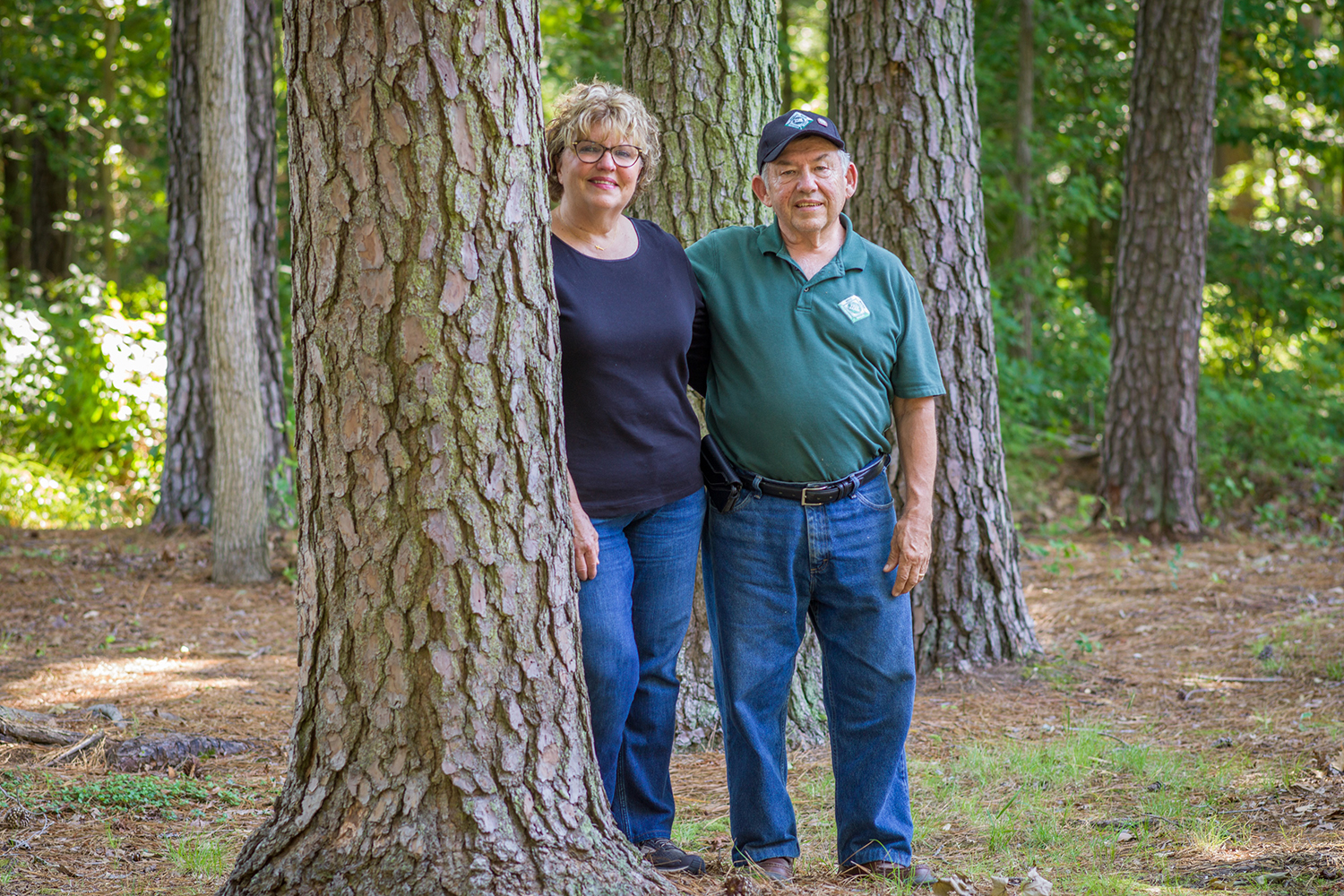 An older woman and man stand close together between large pine trunks in a sunlit forest.