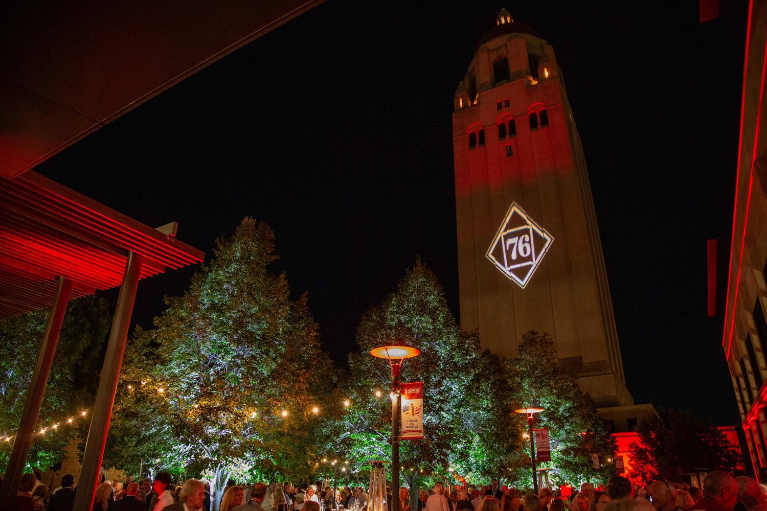 Hoover Tower lit red at night with a glowing class-year emblem, above a crowd gathered under string lights.