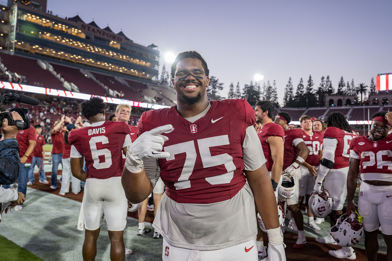 Smiling Stanford football player Braden Marceau-Olayinka, wearing No. 75, gestures on the field after a game as teammates celebrate behind him.