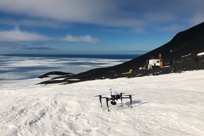 A drone on snow with a small building in the background and water in the distance